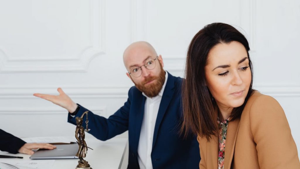 Two professionals in tense discussion beside Lady Justice statue in an office setting.